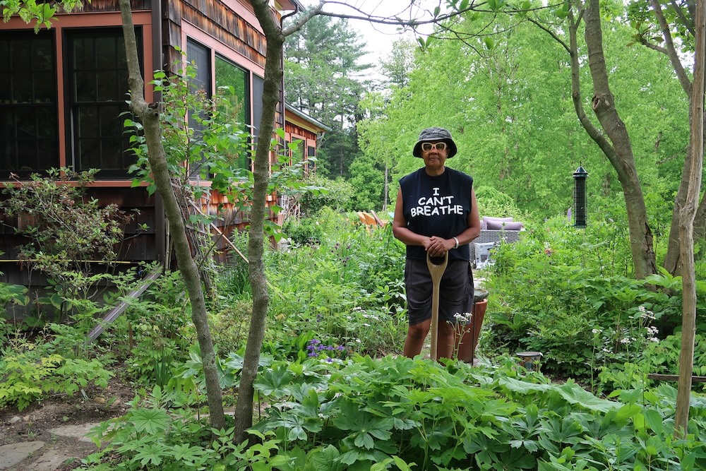 Jamaica Kincaid holding a garden tool in a garden wearing sunglasses and a black t-shirt that reads "I Can't Breathe"